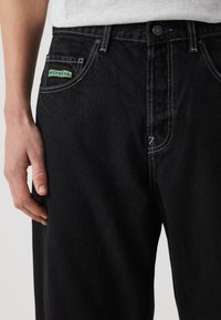 Schwarze Jeans aus Denim mit kontrastierenden weißen Nähten, ausgestattet mit einer Vordertasche mit einem blau-gelben Logopatch. Nahaufnahme des Taillenbereichs.