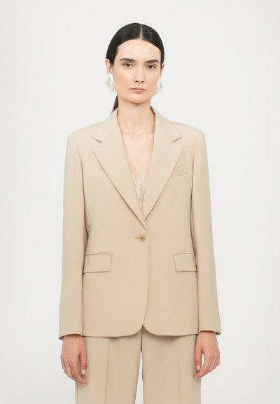 Blazer beige su misura con chiusura a un bottone, colletto a punta e due tasche frontali. Tessuto liscio, vestibilità strutturata, dettagli minimi.