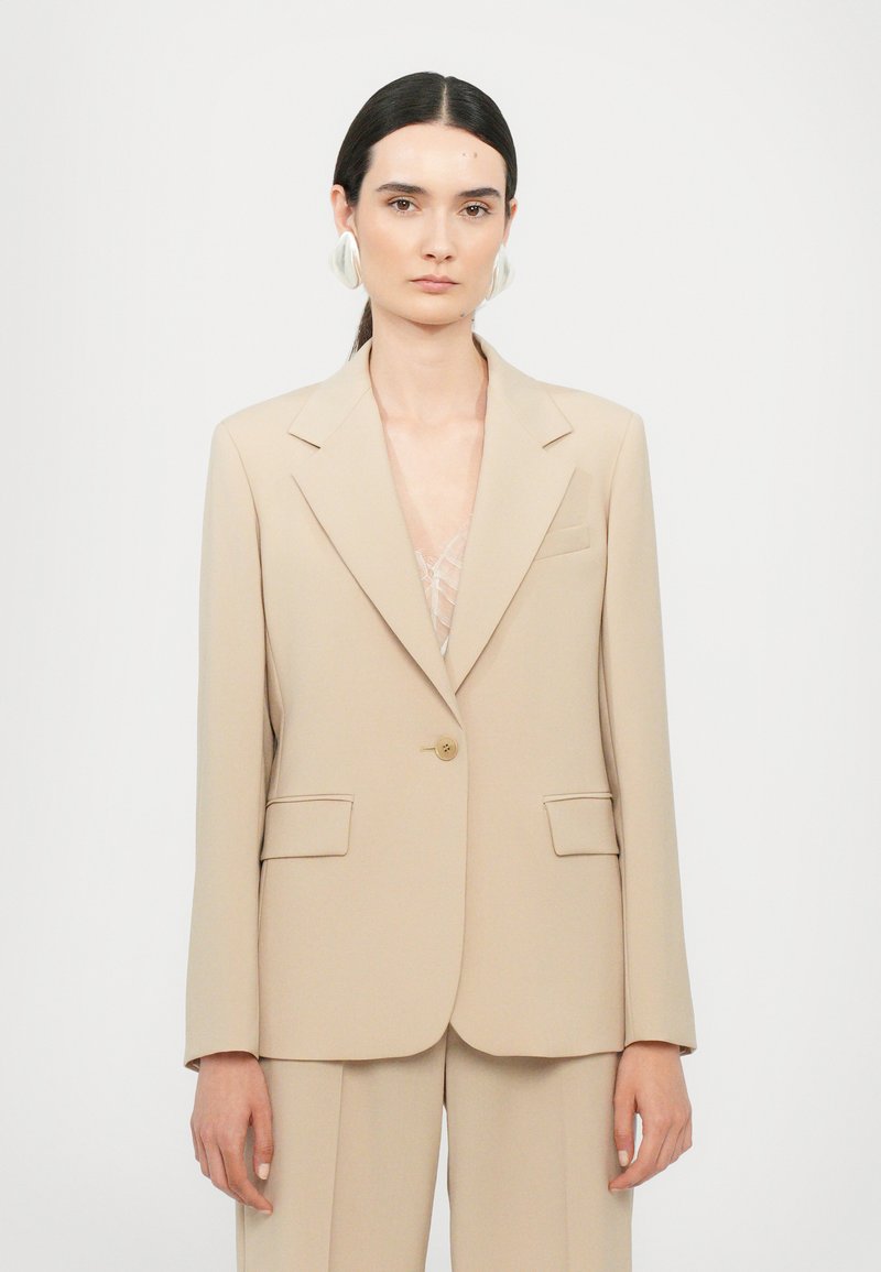 Beige op maat gemaakte blazer met een enkele knoopsluiting, notch-kraag en twee voorzakken. Glad materiaal, gestructureerde pasvorm, minimale details.