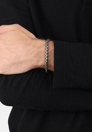 Bracciale in metallo nero con maglie rettangolari, caratterizzato da una superficie testurizzata, indossato su un polso, accompagnato da una maglietta a maniche lunghe nera.