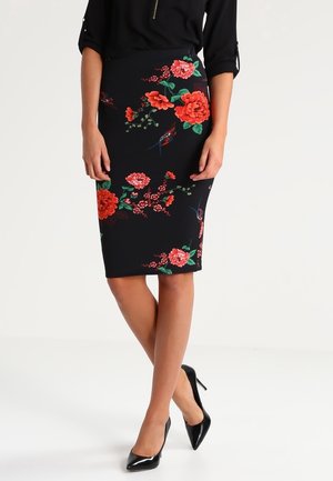 Jupe crayon noire longueur genou avec imprimé floral rouge et oiseaux, portée avec un haut noir et des chaussures à talons hauts noires, jambes croisées.