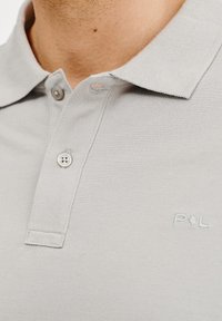 Jasnoszara koszulka polo z teksturowanej tkaniny, z dwoma guzikami, kołnierzykiem oraz małym haftowanym logo na lewej piersi.