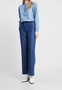 Vaaleansininen denimi-paitapusero, jossa on frillireunus kauluksessa, yhdistettynä korkeavyötäröisiin tummansinisiin leveälahkeisiin farkkuihin ja valkoisiin korkean koron nilkkakorkoihin.