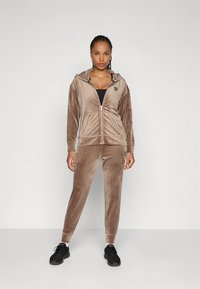 Conjunto de treino em veludo castanho com capuz e fecho éclair, acompanhado por calças justas combinando. Apresenta um acabamento texturado e dois bolsos frontais. Ténis desportivos pretos.