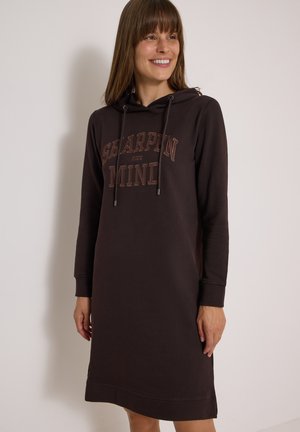Lächelnde Frau, die ein langes schwarzes Hoodie-Kleid mit dem Text "Sharpen Your Mind" trägt, vor einem schlichten hellen Hintergrund stehend.