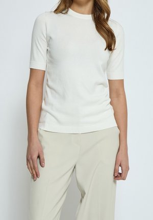 Femme portant un haut en maille blanc ajusté à manches courtes, associé à un pantalon large beige clair, debout devant un fond uni.