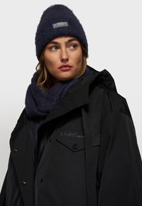 Zwarte waterdichte jas met zijzakken, gecombineerd met een donkerblauwe gebreide beanie en bijpassende textuursjaal. Met merklogo.