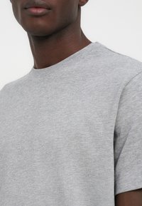 Κοντινό πλάνο ατόμου που φοράει απλό ανοιχτό γκρι μπλουζάκι με στρογγυλή λαιμόκοψη και κοντά μανίκια, σε λευκό φόντο.