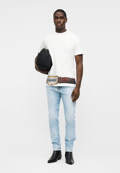 Hombre vistiendo camiseta blanca con diseño estampado de cinturón, jeans azul claro, botas negras, sosteniendo una bolsa negra sobre un fondo liso.