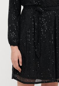 Vestido negro hecho de tela de lentejuelas, con mangas largas, cintura ceñida con un lazo y un dobladillo translúcido.