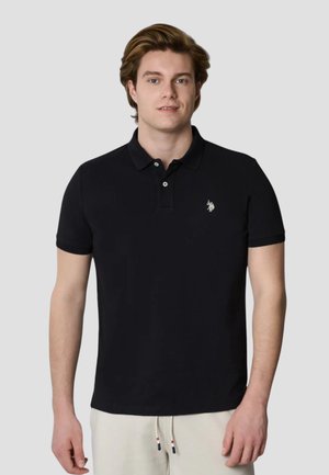 Giovane uomo con capelli castano chiaro che indossa una polo nera con un piccolo logo bianco sul petto sinistro e pantaloni chiari.