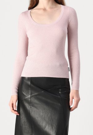 Vrouw draagt een lichtroze geribd, langärmelig top met ronde hals en een hooggetailleerde zwarte leren rok, staand tegen een effen achtergrond.