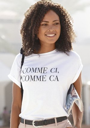 Lächelnde Frau mit lockigem Haar trägt ein weißes Hemd mit der Aufschrift „COMME CI, COMME CA“, hält ein gerolltes Magazin und steht im Freien im weichen Licht.