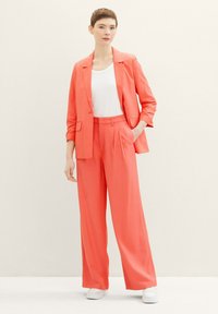 Coral tailored blazer and wide-leg trousers, lightweight fabric, pocket details, paired with a white top and white sneakers. Plain background.