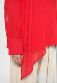 Blouse rouge en tissu transparent avec manches longues, ornée de boutons aux poignets et à la texture lisse et fluide. L'ourlet est asymétrique.