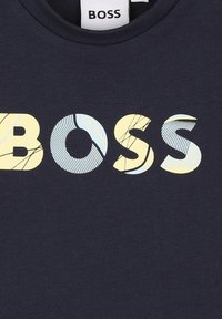 Marineblå T-skjorte med en "BOSS"-logo i gult og lyseblått, som har varierte teksturer og linjemønstre innenfor bokstavene.
