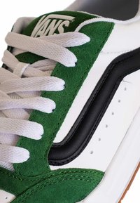 Weißer Sneaker mit grünen Veloursakzenten, schwarzem Designstreifen und weißen Schnürsenkeln. Verfügt über ein strukturiertes Textil und eine Gummisohle.
