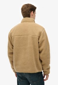 Beige fleecejack met een hoge kraag, zachte textuur en een boxy pasvorm. Heeft een horizontale naad over de rug voor extra detail.