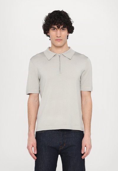 Helles graues kurzärmliges Poloshirt mit einem Reißverschlusskragen, gerippter Textur und lockerer Passform, kombiniert mit dunklen Jeans.
