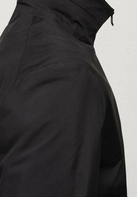 Veste noire avec col montant, texture lisse et fermeture éclair, conçue avec une silhouette angulaire et une esthétique minimaliste.