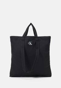 Czarna torba materiałowa z dwoma górnymi uchwytami i małą prostokątną naszywką z logo zawierającym litery "cK". Gładka tekstura, minimalistyczny design.