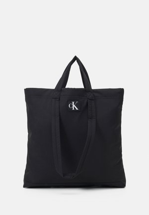 Černá textilní taška se dvěma horními ušima a malou obdélníkovou nášivkou s písmeny "cK." Hladký povrch, minimalistický design.