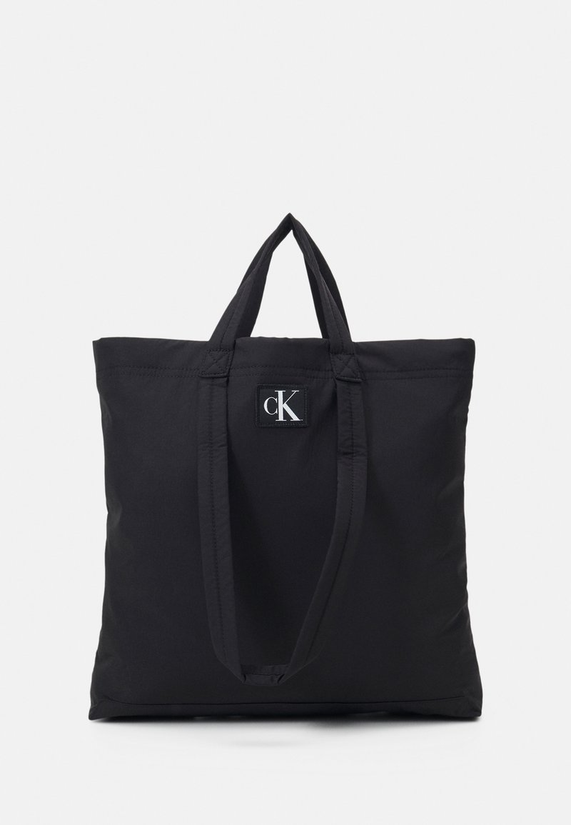 Svart tygväska med två topphandtag och en liten rektangulär logotypatch med bokstäverna "cK." Slät textur, minimalistisk design.