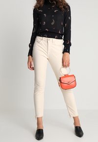 Black long-sleeve top with paisley pattern, cream-colored jeans with frayed hem, holding a small red handbag, and black pointed flats.