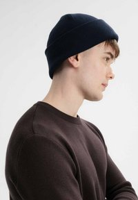 Gebreide marineblauwe beanie met een snug fit, met een ribbelstructuur. Het model draagt een donkerbruine trui en toont een zijaanzicht.