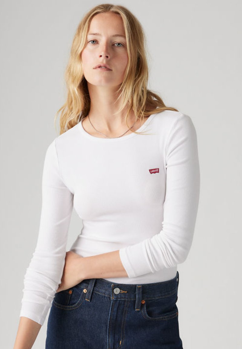 Jeune femme aux cheveux blonds portant un haut blanc à manches longues Levi's et un jean bleu foncé, debout devant un fond uni.