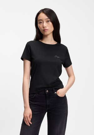 Jeune femme aux longs cheveux foncés portant un t-shirt noir et un jean noir, une main dans la poche, debout devant un fond blanc.