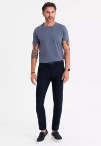 Šedé tričko s krátkým rukávem, sladěné s tmavě modrými slim-fit kalhotami a tmavými slip-on botami. Na rukou má tetování a hodinky na zápěstí.