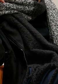 Svart jacka i ullblandning med en strukturerad, mörkgrå sjal som har ett ljusgrått mönster, hängande löst över axlarna.