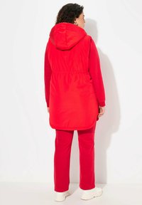 Roten Kapuzenweste mit elastischem Bund und seitlichen Schlitzen, kombiniert mit roten Hosen und weißen Sneakers, vor einem weißen Hintergrund präsentiert.