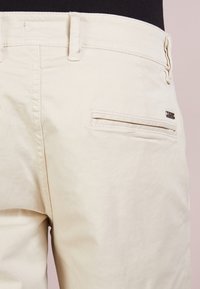 Pantalon en coton beige clair avec devant plat, poche arrière à ouverture horizontale et passants pour ceinture. Texture lisse avec peu de détails.