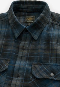 Cămașă din flanel în nuanțe de albastru închis, negru și un model tartan maro. Are o guleră tip spread, nasturi negri și două buzunare la piept.