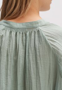 Blusa verde chiaro con una vestibilità rilassata, caratterizzata da pieghe e un tessuto morbido e testurizzato. Il dettaglio arricciato sulla scollatura ne esalta la forma.