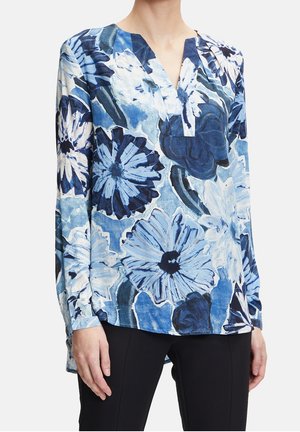 Frau trägt eine langärmlige Bluse mit großem blau-weißem Blumenmuster und V-Ausschnitt, kombiniert mit schwarzen Hosen.