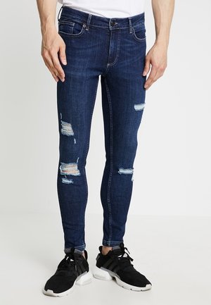Jean slim bleu foncé déchiré porté avec des baskets noires et blanches sur une personne debout sur un fond uni.