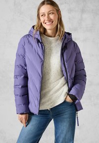Lila puffjacka med huva har ett chevron-quiltmönster och svarta dragkedjeaccenter, bärs över en ljusgrå texturerad tröja och blå jeans.