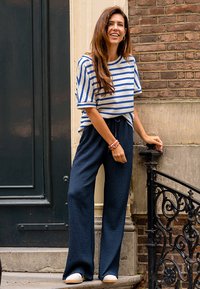 Gestreepte gebreide shirt in wit en blauw. Loszittende marineblauwe textuurbroek. Staand op trappen bij een decoratieve zwarte reling.