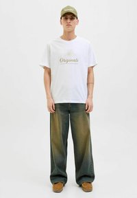 Camiseta de algodón blanca con "Originals" impreso en dorado. Jeans de corte ancho oversize en verde oscuro. Zapatos marrones sin cordones. Gorra verde oliva.
