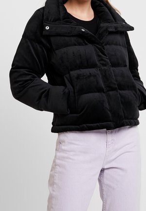 Giacca nera imbottita con una consistenza morbida, caratterizzata da un alto colletto, bottoni a pressione e due tasche frontali; indossata con pantaloni di un leggero colore viola.