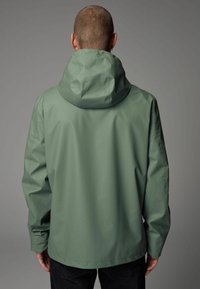 Jachetă verde impermeabilă cu glugă, având o textură netedă și o croială lejeră. Partea din spate prezintă cusături simple și fără prinderi vizibile.