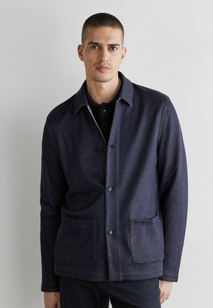 Mand med kort hår iført en mørkeblå knaplukket jakke med to forlommer, stående mod en ensformet hvid væg.