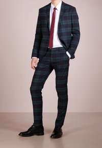 Donker geruit pak met groene en marineblauwe patronen, een enkel-breasted jas, slim-fit broek, aangevuld met een kastanjebruine stropdas en zwarte nette schoenen.