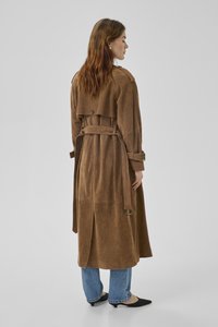 Vrouw met lang bruin haar draagt een suède trenchcoat met ceintuur, een blauwe spijkerbroek en zwarte kittenheels, en staat met haar rug naar de camera op een effen achtergrond.