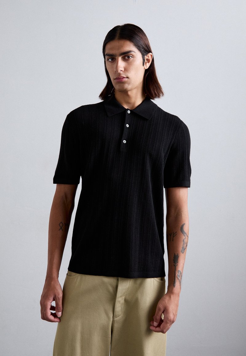 Polo din tricot negru, cu guler, sistem de închidere cu trei butoane, mâneci scurte și textură cu dungi verticale.