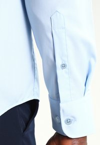 Camisa de manga larga azul claro, hecha de tejido suave. Presenta un puño con puntas y dos botones, y un dobladillo recto.