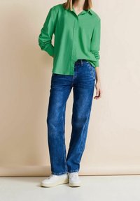 Grön skjorta med knappar och långa ärmar, tillsammans med blå raka jeans och vita sneakers på en neutral bakgrund.